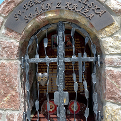 Kovaný památník svatých s atributy na mřížích. Sv. Terezie z Avily - srdce s nápisem IHS, Sv. Jan od Kříže - kříž