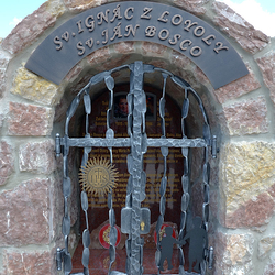 Kovaný památník svatých s atributy na mřížích. Sv. Ignác z Loyoly - nápis IHS, Sv. Ján Bosco - děti